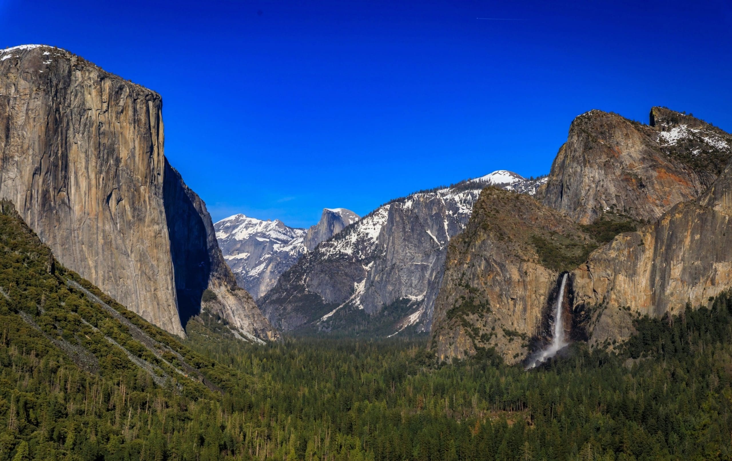Majestic mountain landscape with cliffs, snowy peaks, a forested valley, and a waterfall beneath a clear blue sky.