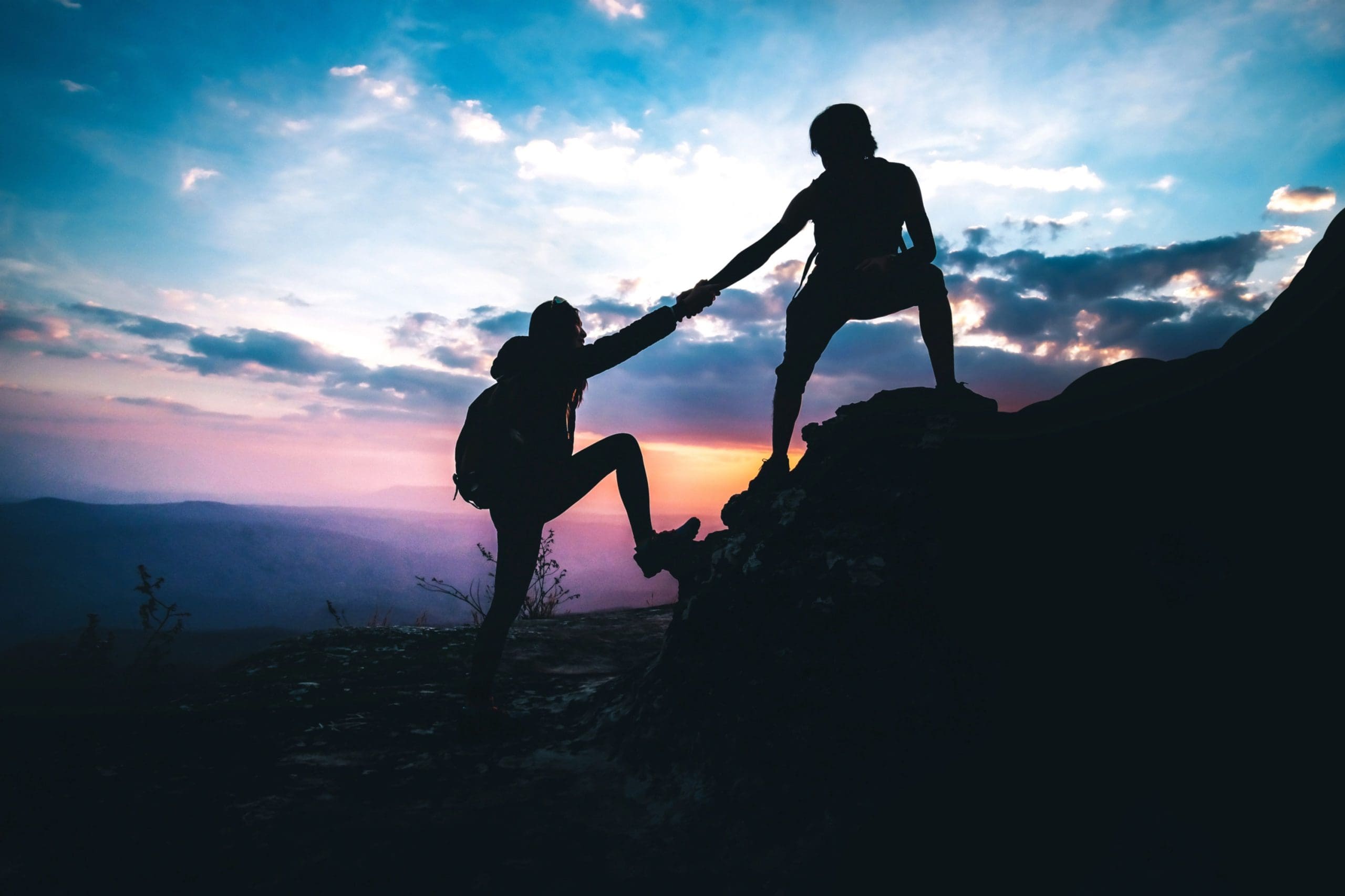 Silhouette of one hiker helping another up a rocky peak at sunrise, symbolising teamwork and support.