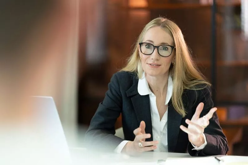 Woman wearing glasses speaking and gesturing with her hands during a meeting in a professional setting.