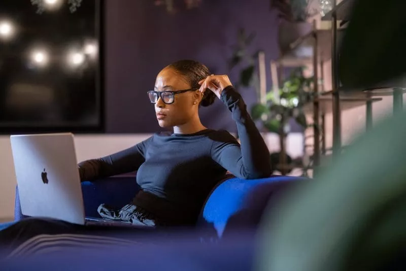 Woman wearing glasses sitting on a sofa and working on a laptop in a softly lit room with plants in the background.