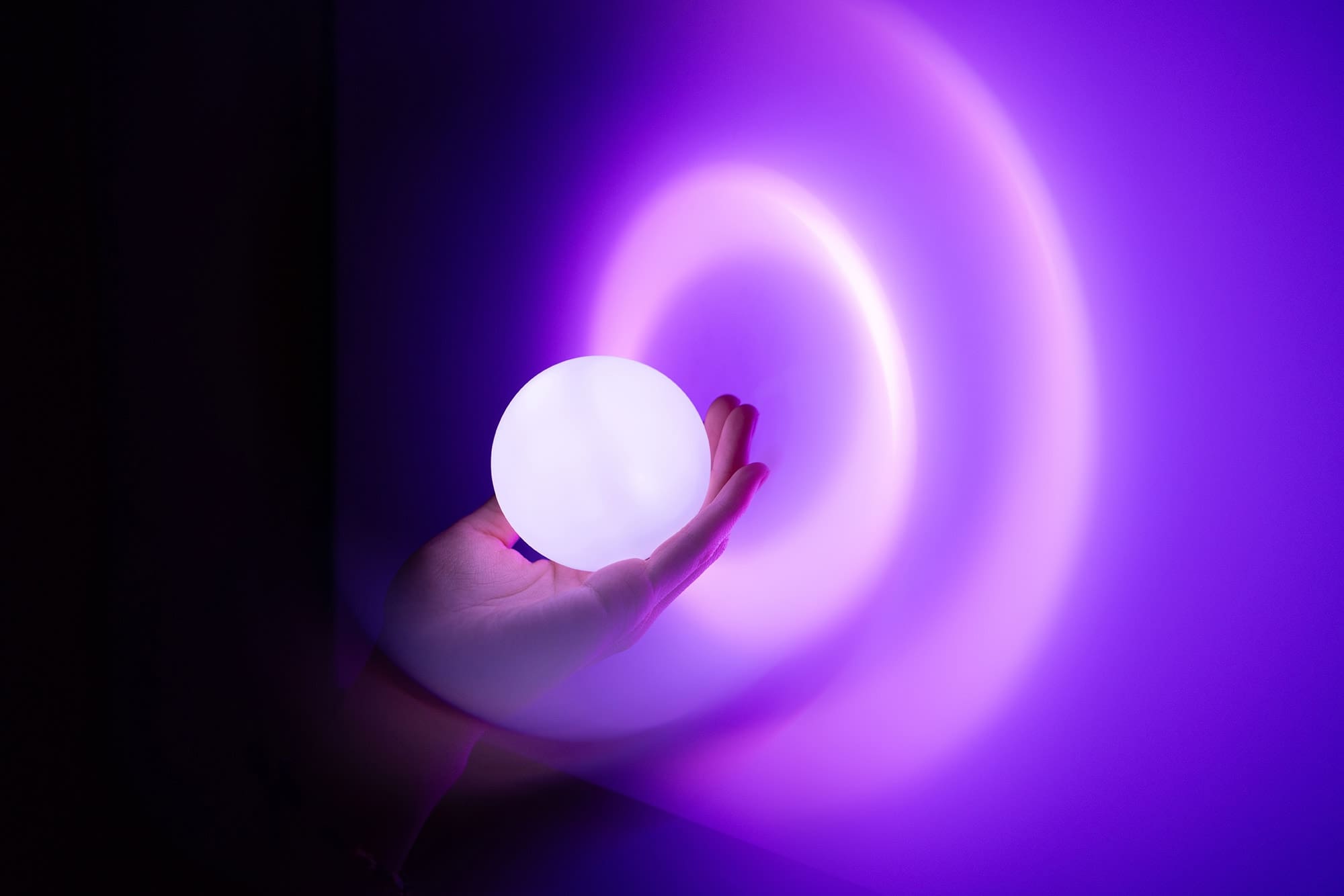 A hand holding a glowing white orb against a dark background, casting concentric rings of purple light onto the wall.