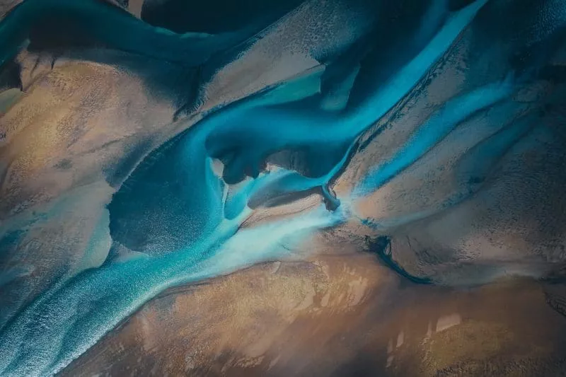 Aerial view of a winding river delta with vibrant turquoise and deep blue water channels flowing through brown and tan sandy terrain.