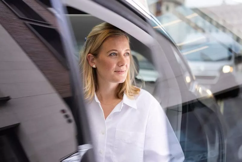 Person in a white shirt sitting in the back seat of a car, looking out the open door with a calm expression.