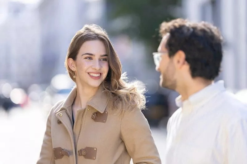 Two people talking outdoors on a sunny day, with one person smiling brightly while wearing a tan coat.