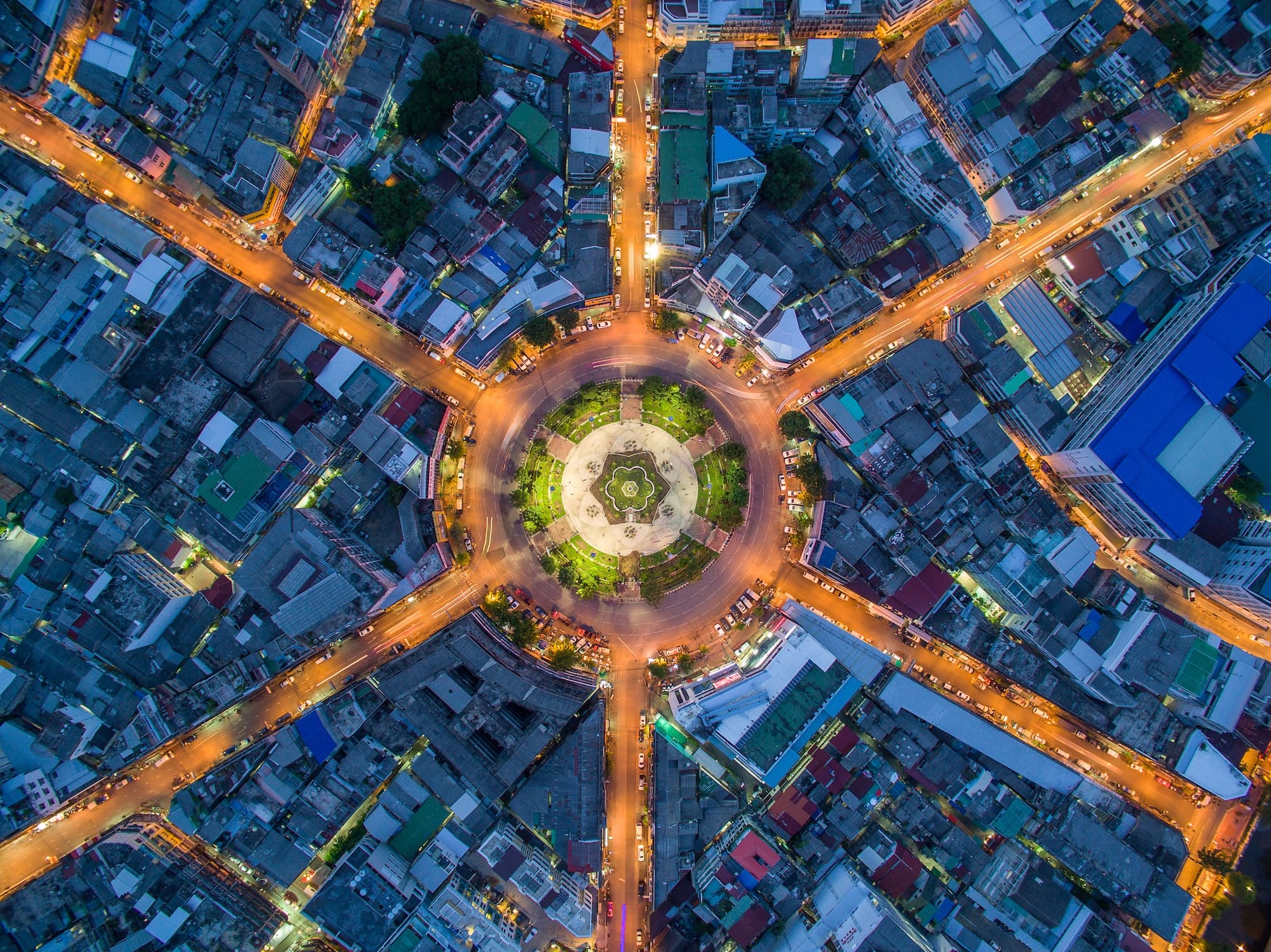 Aerial view of a brightly lit roundabout in a densely packed urban area at night, with streets radiating outward like spokes and buildings tightly surrounding the circle.