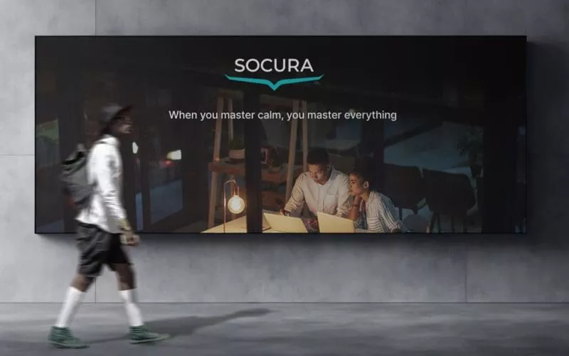 Person walking past a wall-mounted digital billboard showing two people working at laptops under warm lighting, with the text “SOCURA — When you master calm, you master everything.”