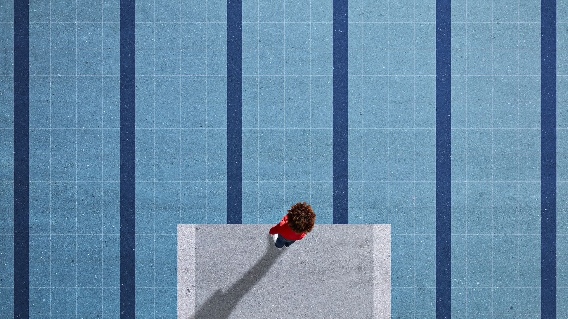Aerial view of a person standing alone at the edge of a concrete platform, facing an empty blue swimming pool with lane markings, creating a sense of pause and anticipation.