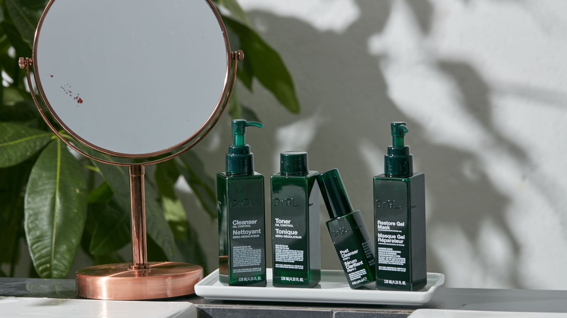 DrGL skincare products displayed on a white tray beside a copper vanity mirror, including cleanser, toner, serum, and restore gel mask in dark green bottles, set against a bright, leafy indoor background.