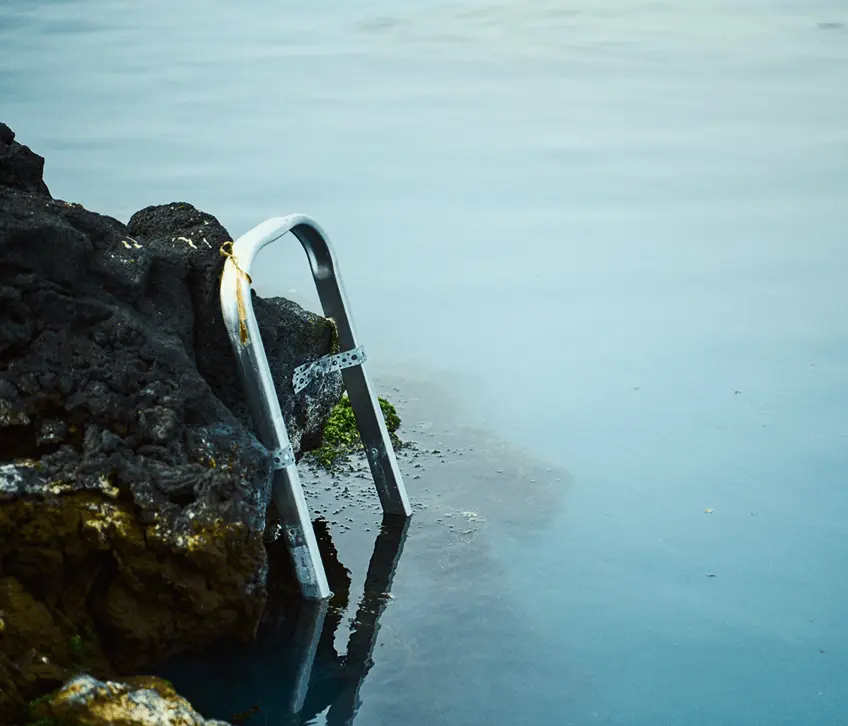 A candid editorial-style photograph showing a group of Icelandic workers and community members engaged around HS Orka’s geothermal site. The scene includes people in workwear interacting with natural and industrial elements—such as rugged lava rock landscapes, steam vents, power plant structures, and geothermal equipment—capturing dramatic Icelandic light and weather conditions to reflect the human side of the energy company’s operations. This photography style is part of HS Orka’s brand work emphasizing real people and place within its renewable energy story.
