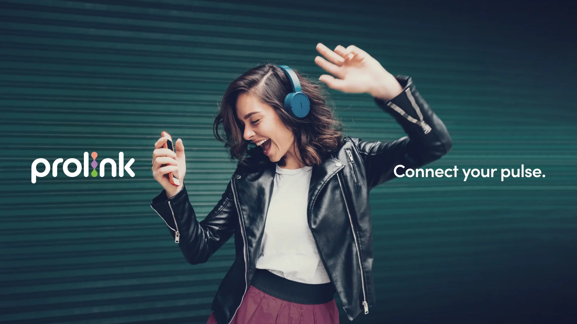 A woman wearing blue over-ear headphones and a black leather jacket dances joyfully while looking at her smartphone. She stands in front of a green corrugated metal wall. On the left is the “prolink” logo, and on the right the text reads, “Connect your pulse.”