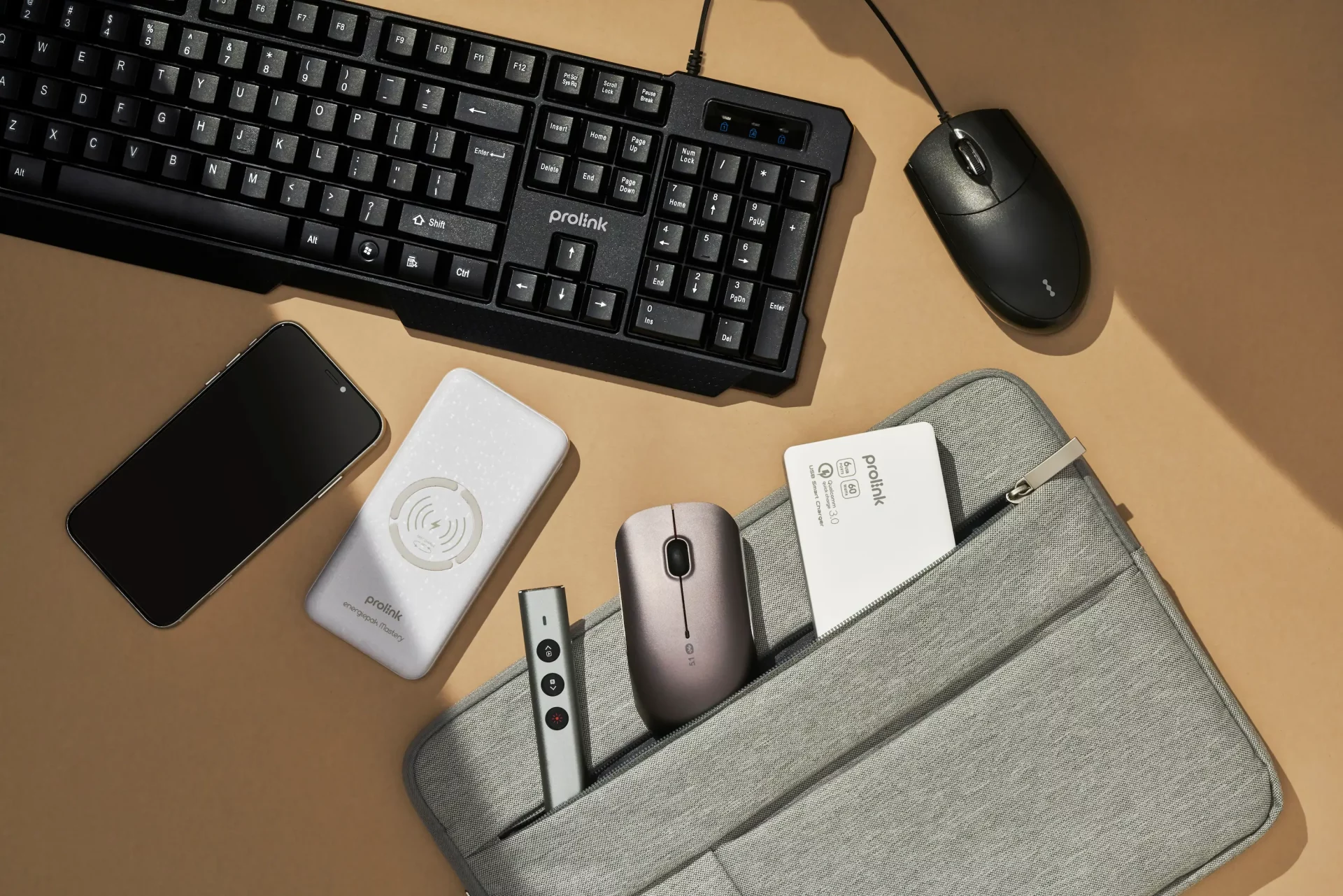 Top-down view of a tidy workspace featuring Prolink accessories: a smartphone, a white wireless power bank, a slim presentation remote, and a wireless mouse partially tucked into a grey fabric laptop sleeve. A Prolink keyboard and mouse sit at the top of the desk, emphasizing a coordinated, modern work-from-anywhere setup.