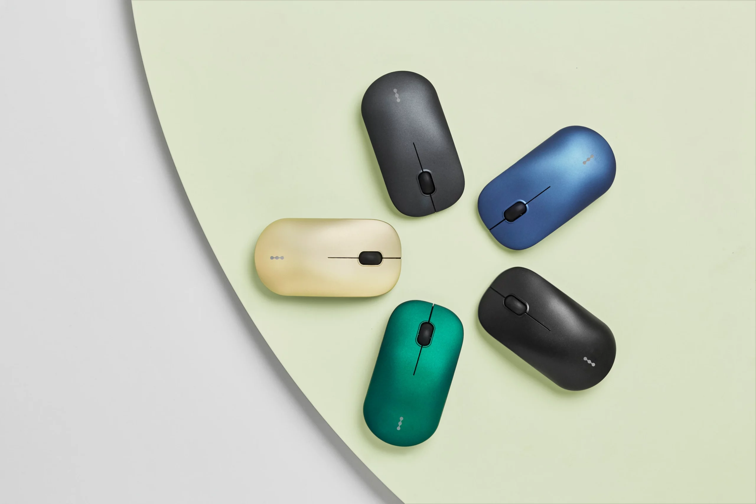 Top-down view of five compact, pebble-shaped devices in black, blue, green, yellow, and dark grey, arranged in a loose circular pattern on a soft, two-tone background with curved edges, highlighting color variety and minimalist design.