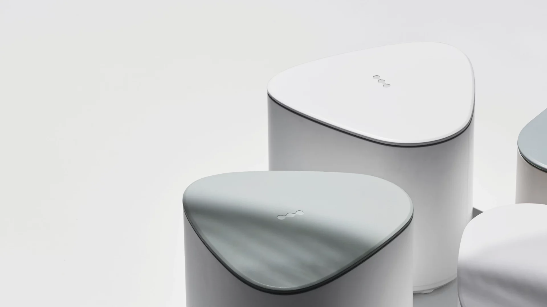 Close-up of three minimalist, rounded triangular devices with smooth matte finishes in light grey and pale green, arranged on a neutral background. Each device has a small abstract logo centered on the top surface, emphasizing a clean, modern product design.