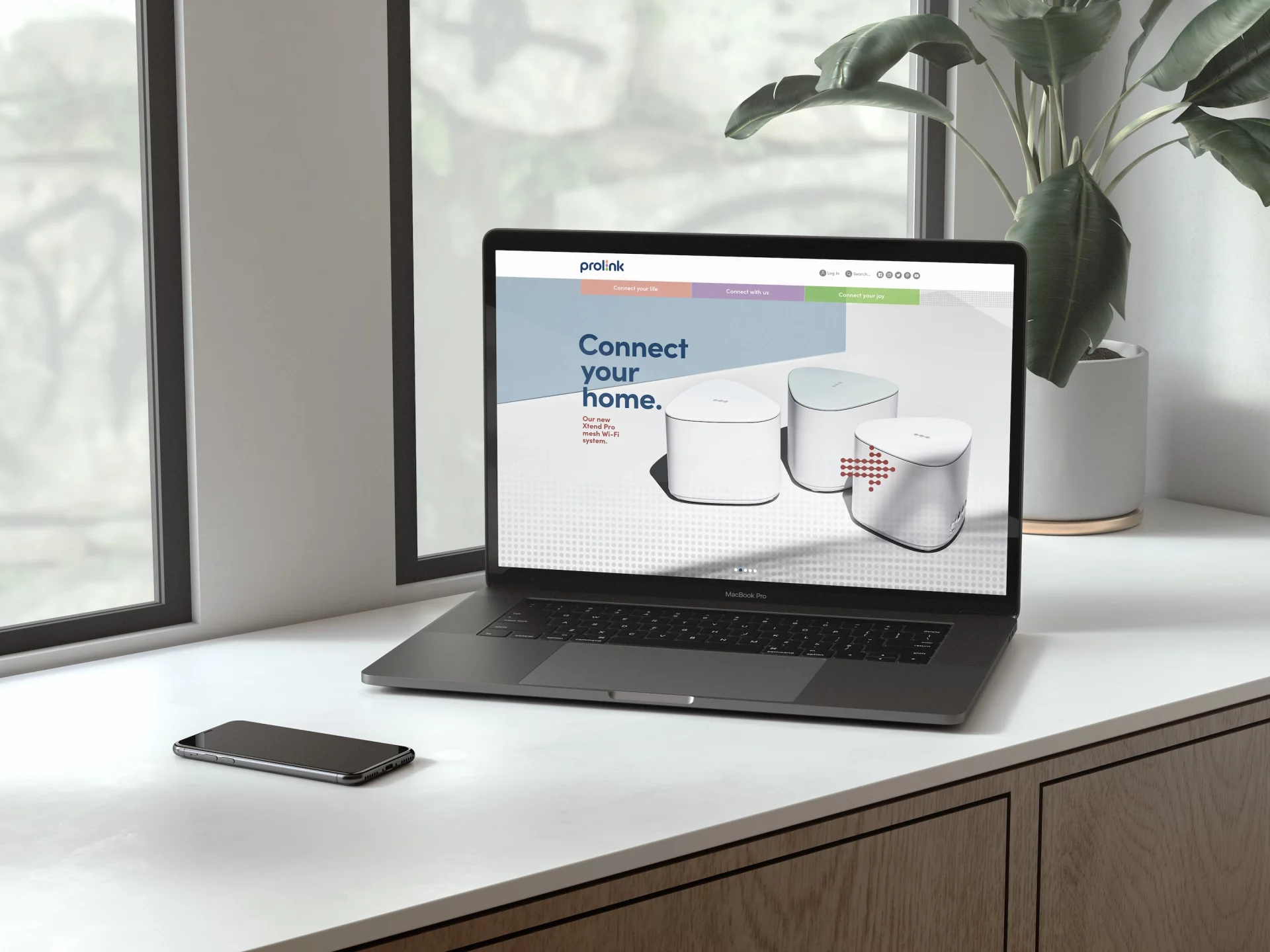 A laptop on a clean desk displays the Prolink website featuring a “Connect your home” banner and images of sleek mesh Wi-Fi devices. A smartphone rests beside the laptop, with natural light from a window and a potted plant creating a calm, modern workspace atmosphere.