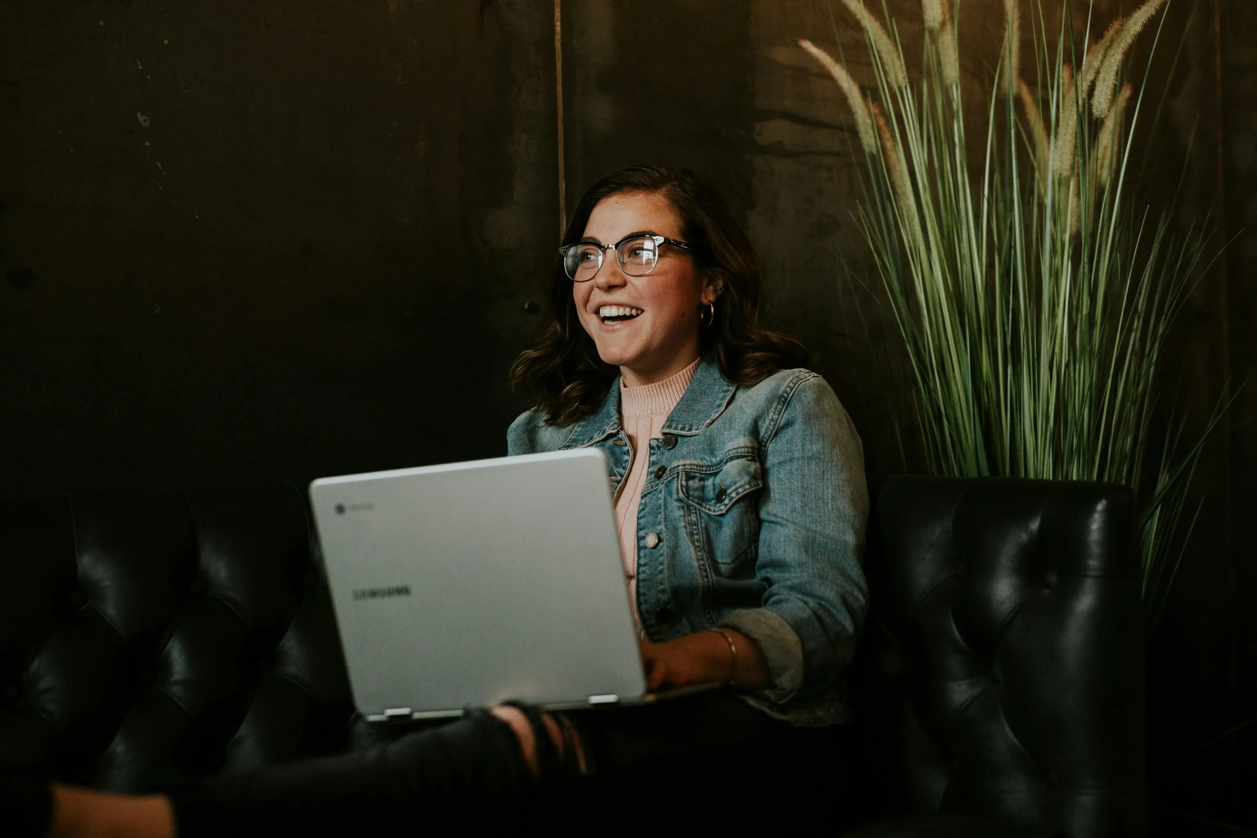A smiling woman wearing glasses and a denim jacket sits on a leather sofa using a laptop in a modern, dimly lit workspace with tall decorative plants in the background.