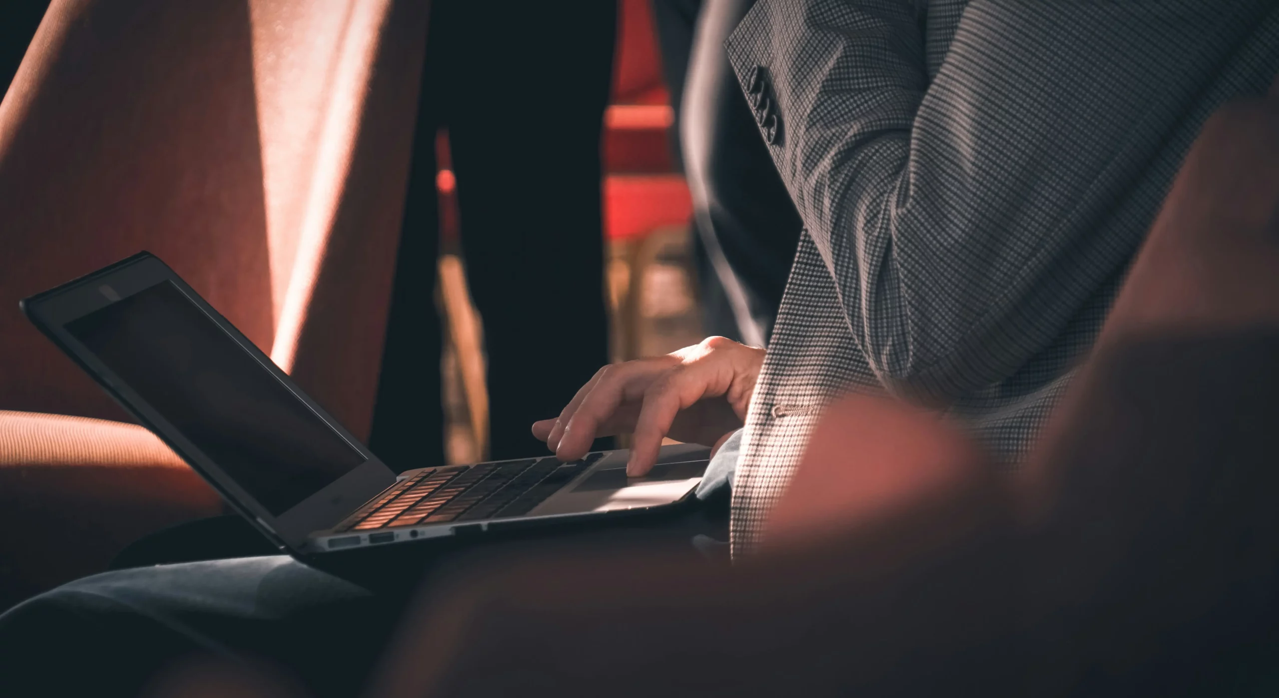 A business professional using a laptop while seated in a meeting or conference setting, with warm lighting and blurred figures in the background.
