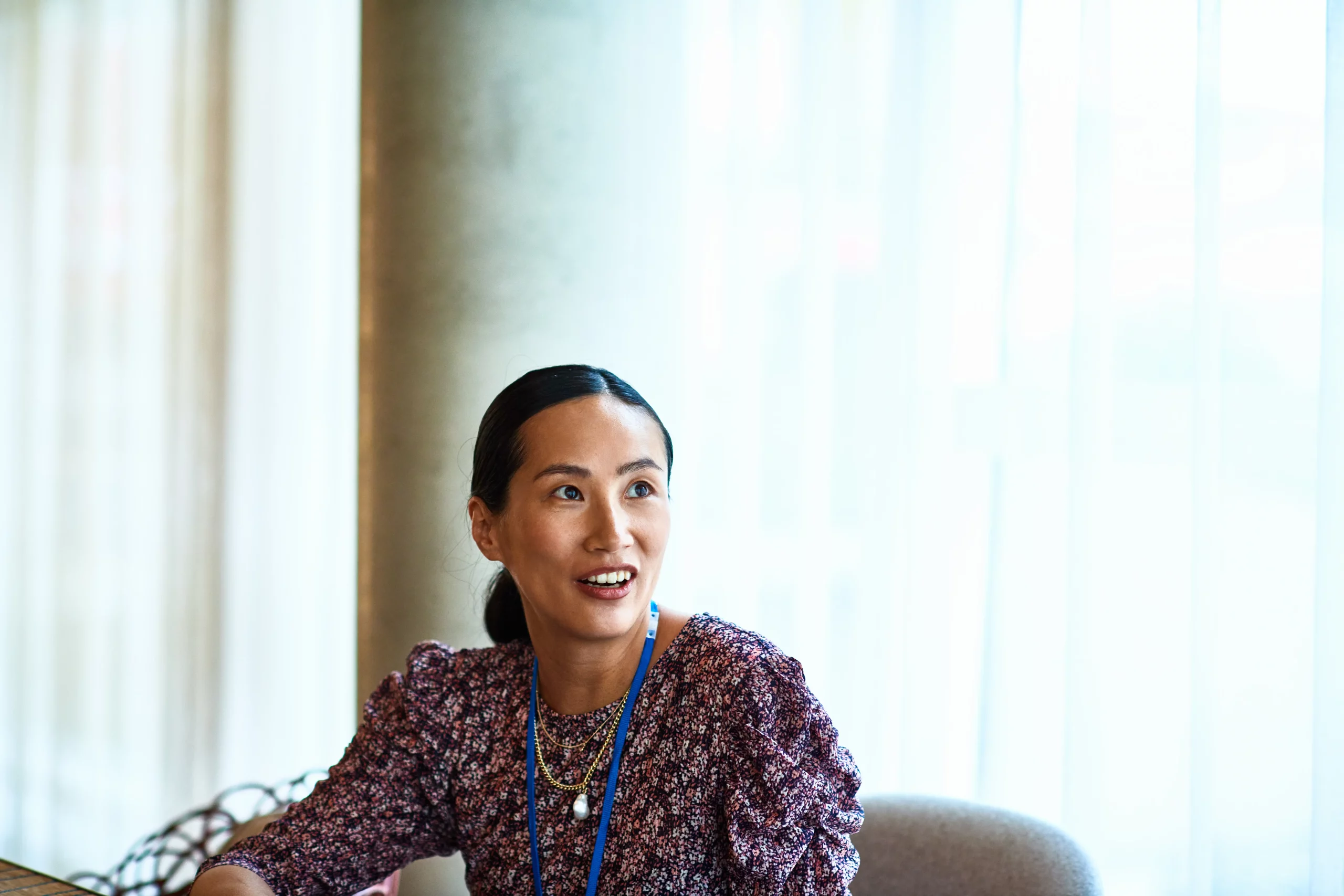 A woman wearing a patterned blouse and a blue lanyard sits at a table indoors, looking to the side while speaking, with soft natural light coming through large windows behind her.