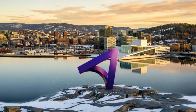 A modern city skyline at sunset reflected in calm harbor water, with snowy hills in the background. In the foreground, a large purple abstract sculpture shaped like a curved, angular “7” stands on a snow-dusted rocky outcrop. To the right, a striking white, sloped-roof building sits along the waterfront, and boats are docked in a marina nearby.