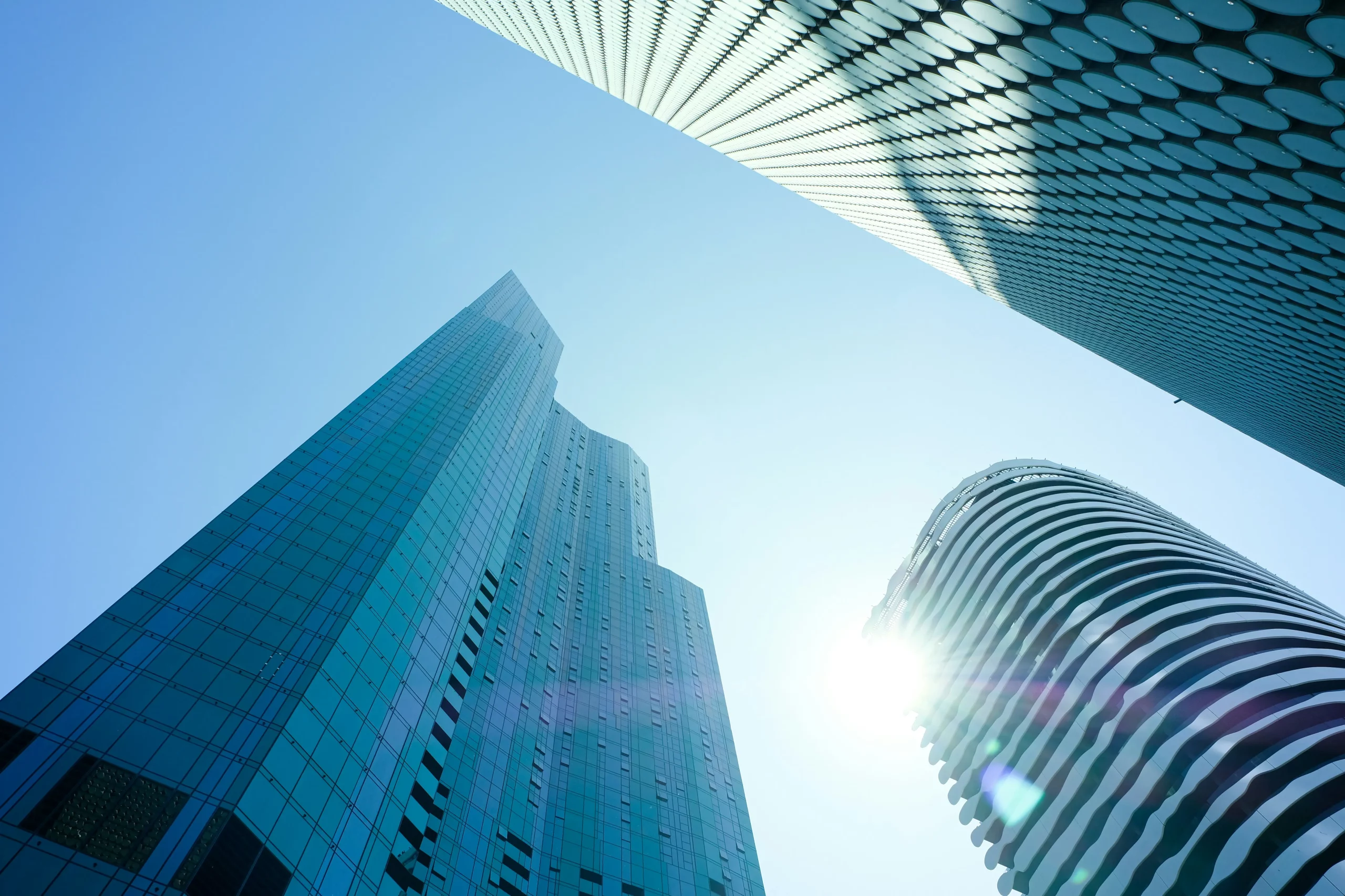 Glass skyscrapers viewed from below against a clear blue sky, with sunlight reflecting off modern curved and geometric facades.