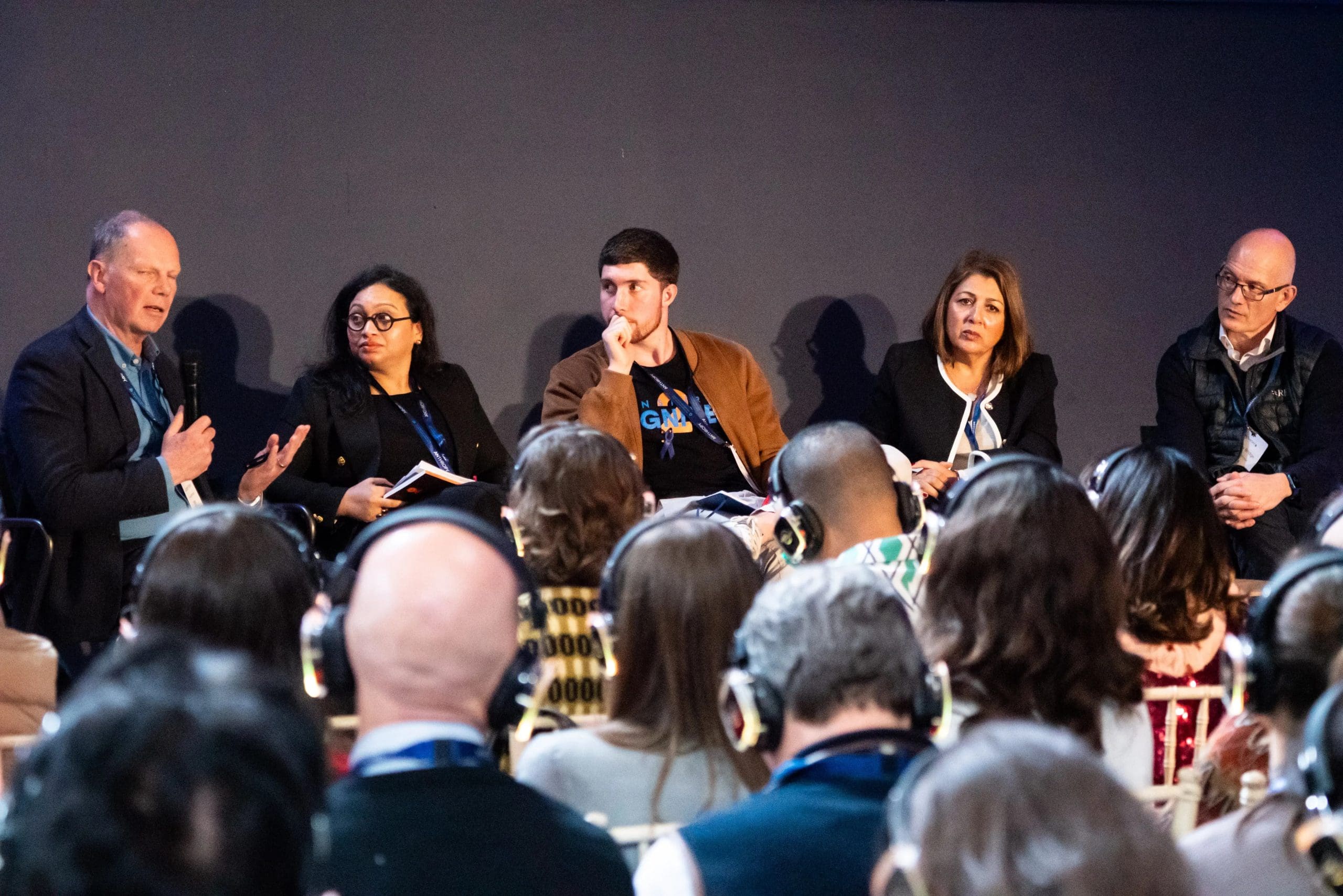 Panel discussion on stage with five speakers seated in a row, while audience members wearing headphones listen during a conference session.