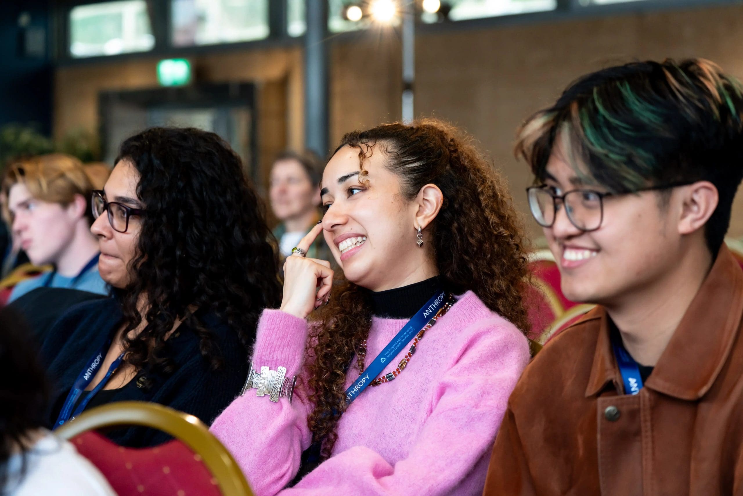 Three conference attendees wearing event lanyards sit in the audience smiling and laughing while listening to a speaker.