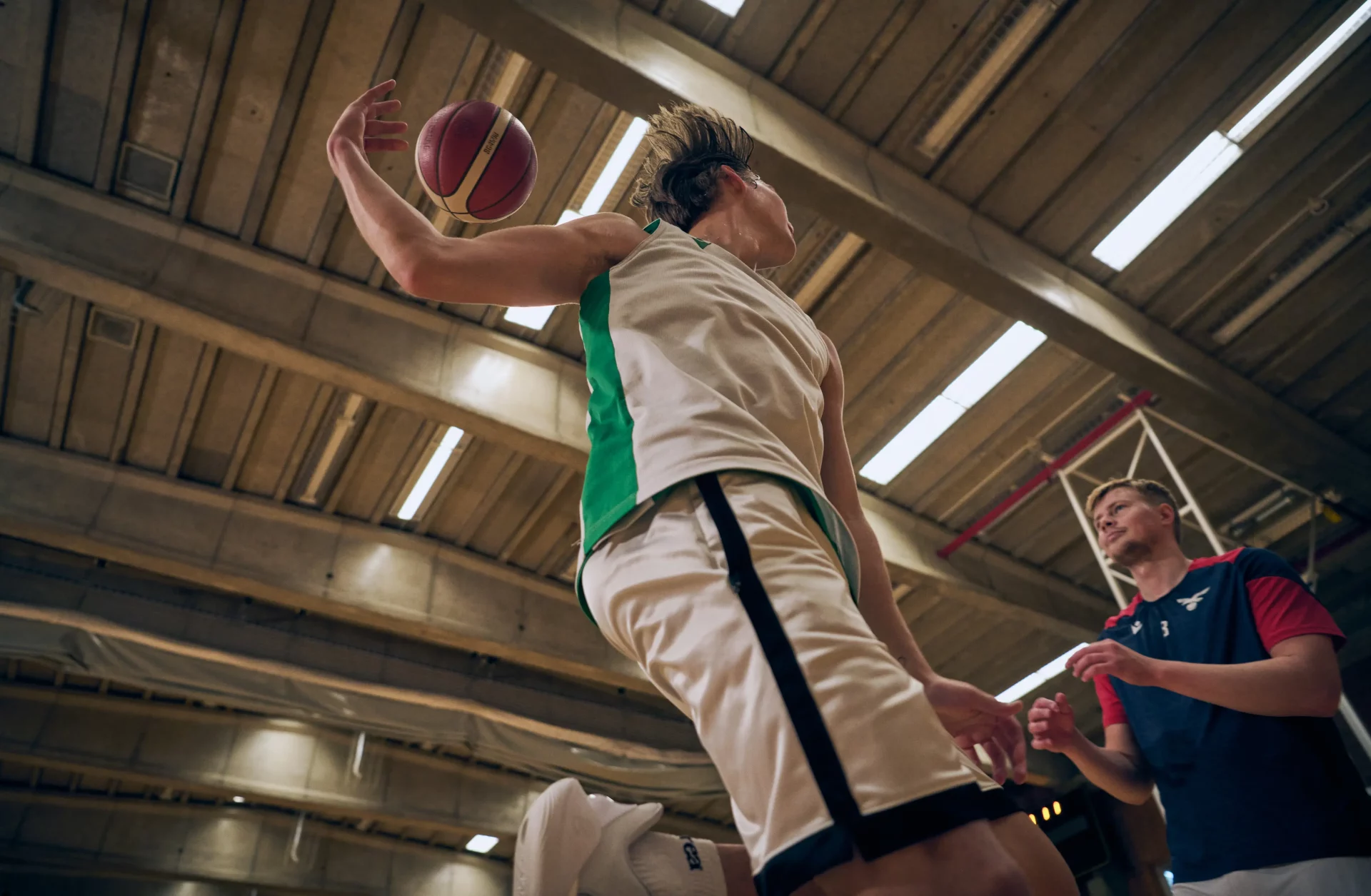 Low-angle view of a basketball player in a white and green uniform mid-jump holding a basketball overhead, with another player watching, inside an indoor gymnasium.