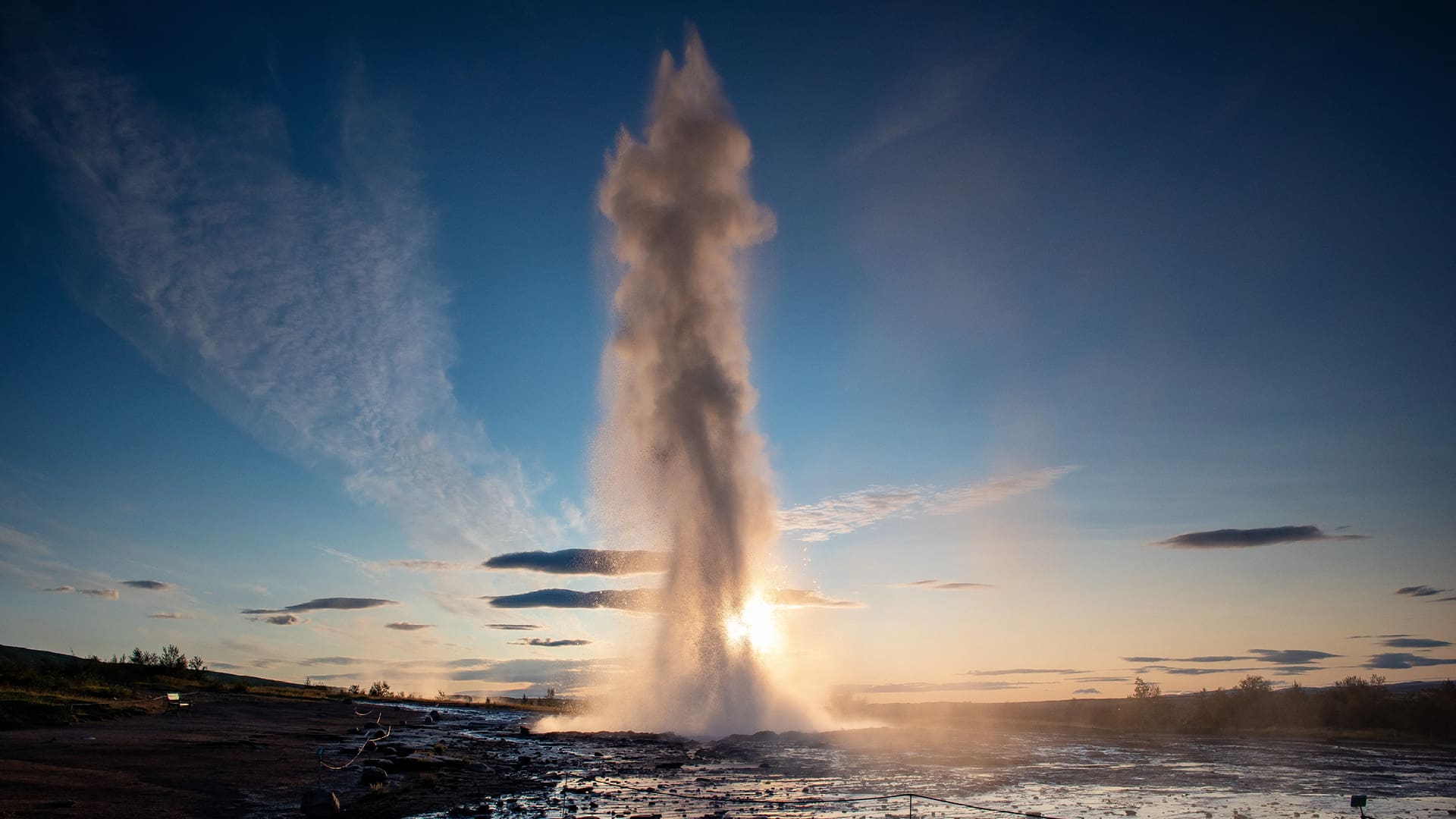 A geyser erupts from the ground, shooting a tall column of water and steam into the air against a clear blue sky. The low sun behind it creates a glowing halo and backlights the spray. The surrounding landscape is flat and rocky with shallow water pools, and a few clouds stretch across the horizon.
