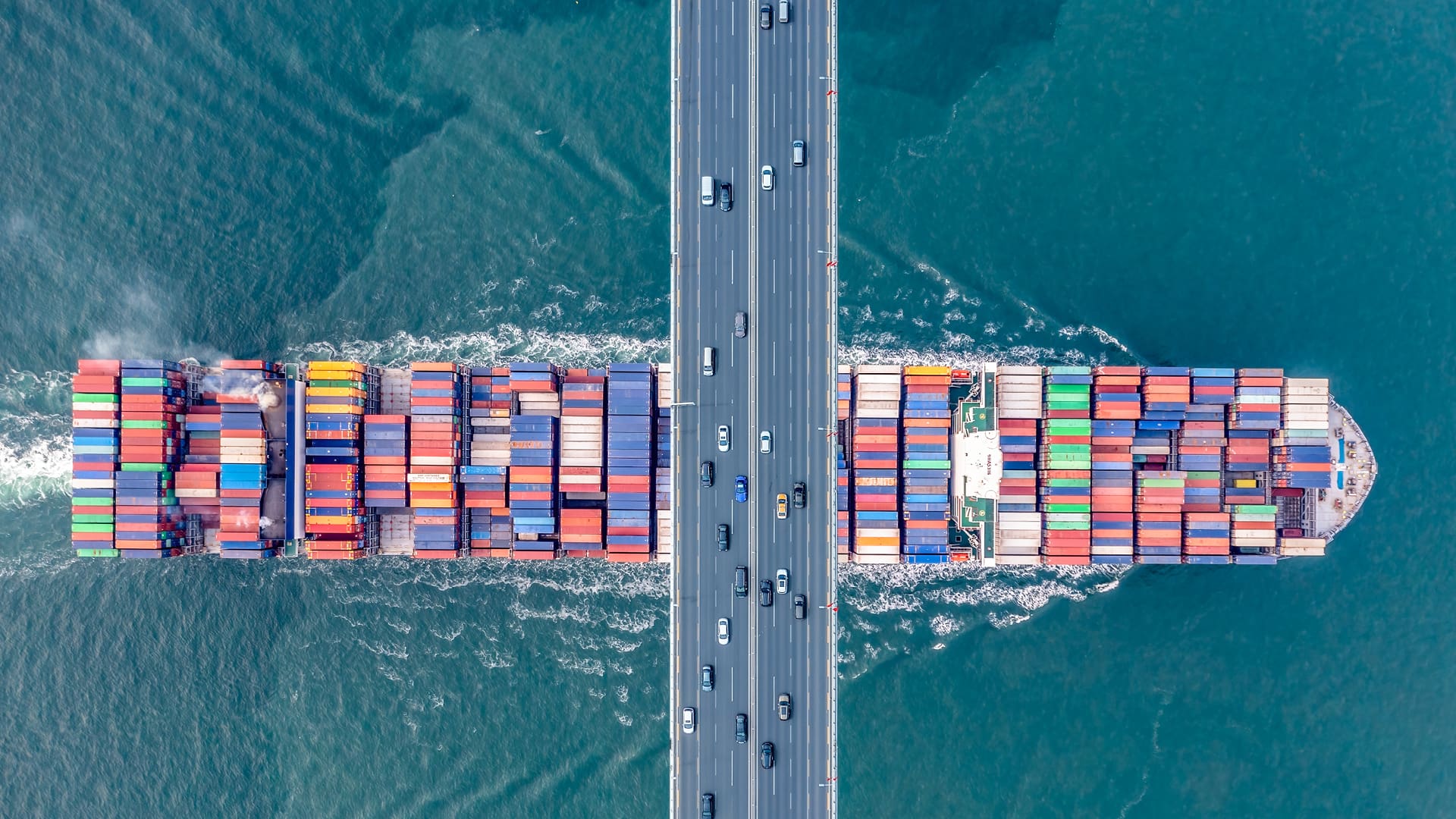 Aerial view of a container ship stacked with colorful shipping containers passing beneath a multi-lane highway bridge, with cars traveling overhead and turquoise water surrounding the vessel.