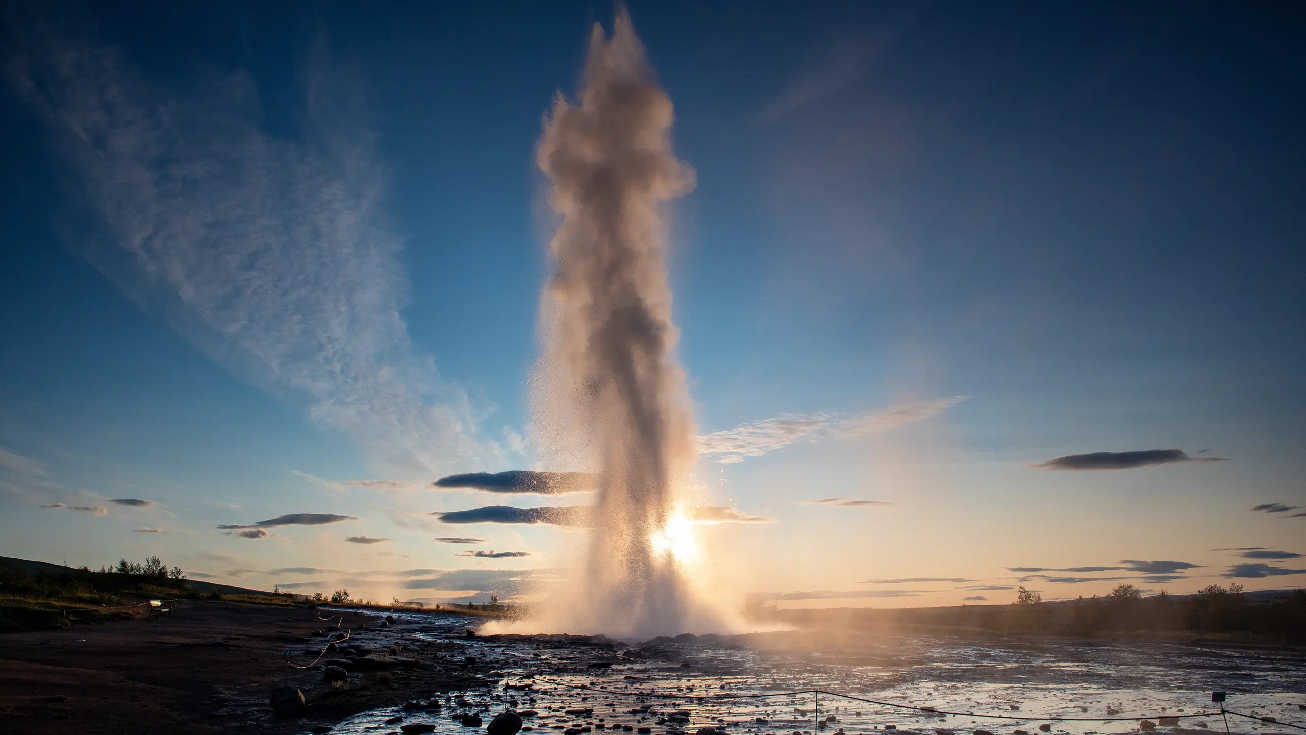 A tall geyser erupts, sending a column of water and steam high into the air at sunset. The sun glows behind the plume, illuminating the mist, with a rocky, wet landscape and scattered clouds in the sky.