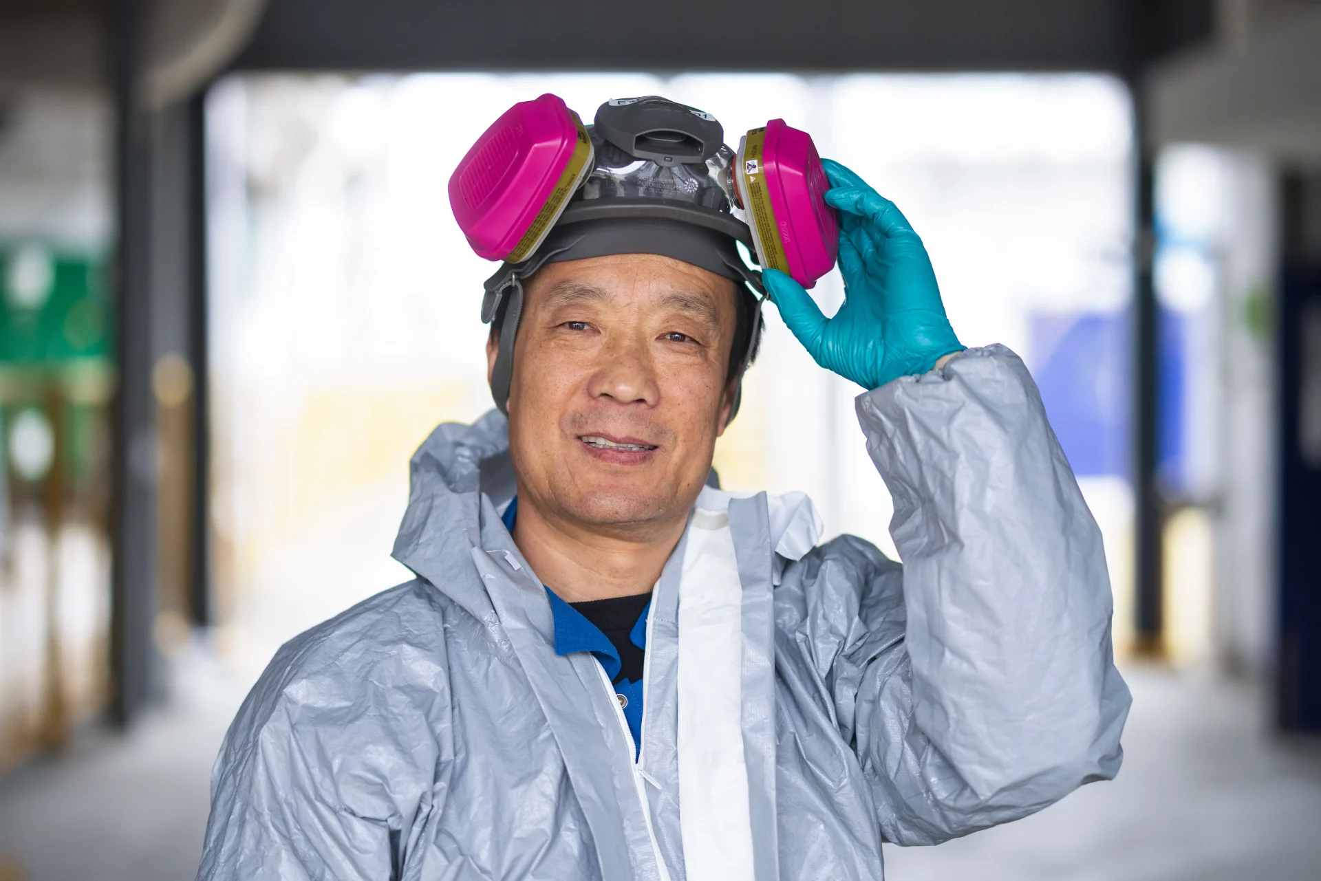 OneSubsea employee wearing a gray protective suit and teal gloves, lifting a respirator mask with pink filters on his head, standing indoors in an industrial setting.