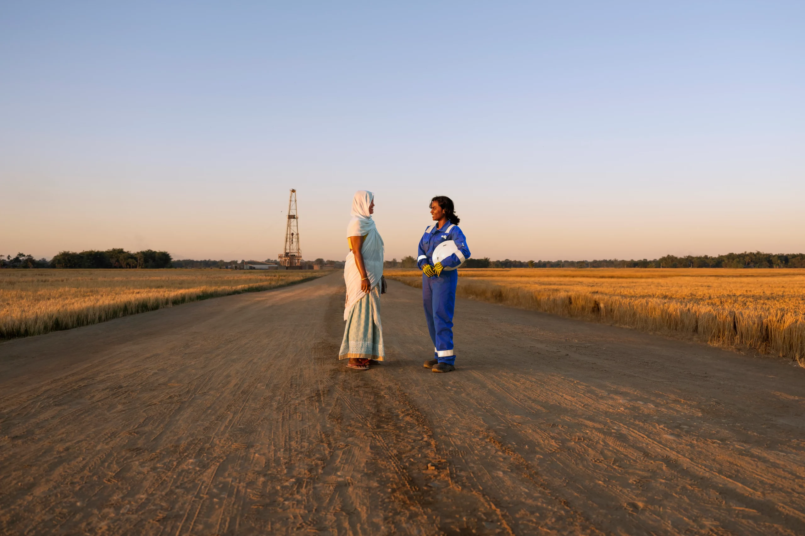 Two women standing and talking on a dirt road between golden fields at sunset, with one wearing traditional clothing and the other in blue industrial coveralls holding a white hard hat, and an oil rig visible in the distance.