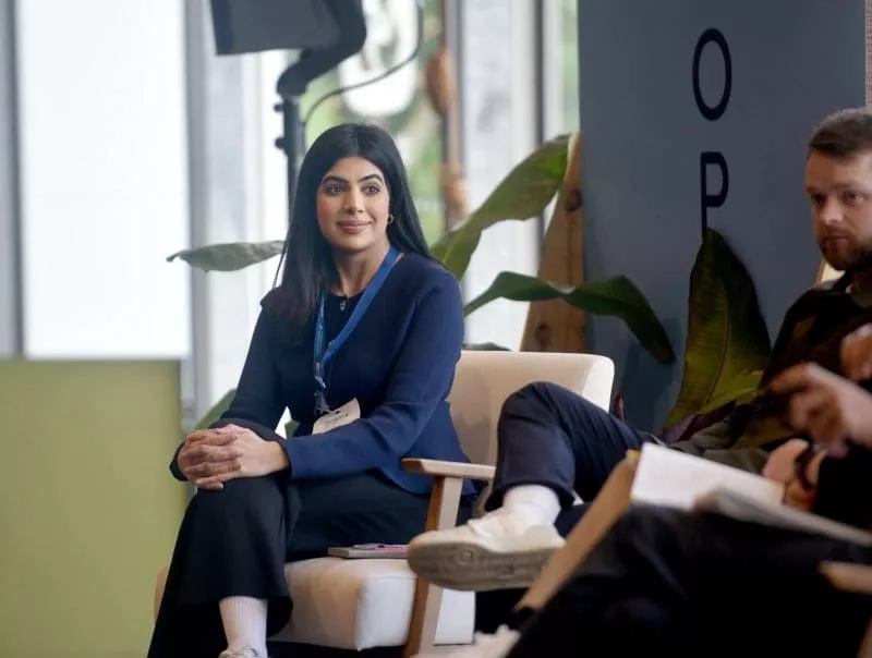 Woman wearing a conference lanyard seated in an armchair on a panel discussion stage, listening attentively with hands clasped, with other panelists partially visible beside her.