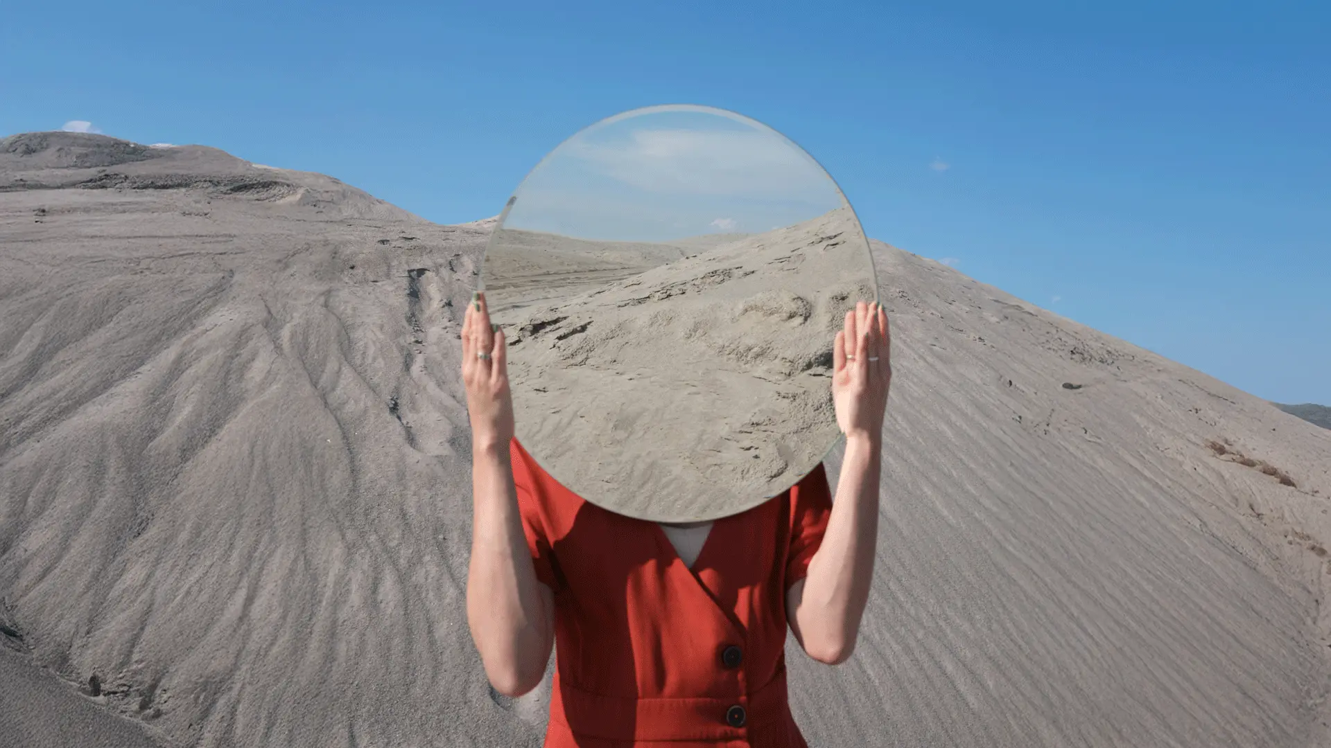Person in a red dress standing in a barren, sandy landscape holds a large round mirror in front of their face, reflecting the surrounding hills and blue sky so that their face is completely hidden.