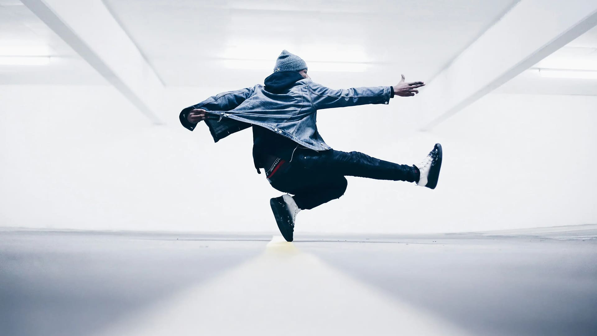 Person in a denim jacket and beanie performing a mid-air dance move in a bright, empty parking garage.