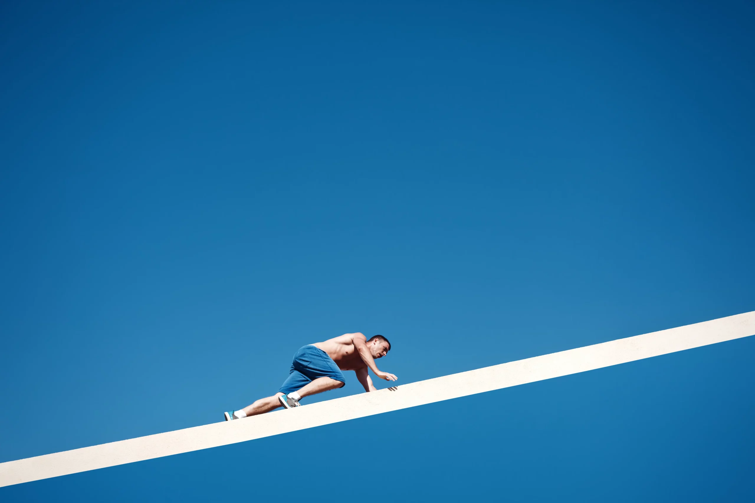 Shirtless man in blue shorts and sneakers balancing on a narrow white beam against a clear blue sky.