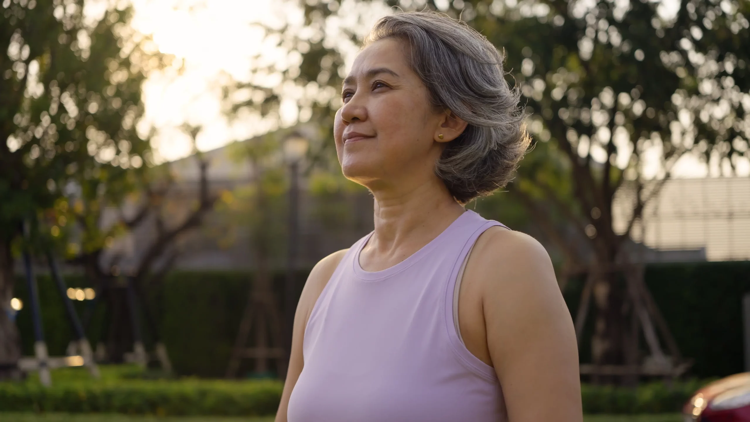 Middle-aged woman with gray hair standing outdoors in a park at sunrise, looking ahead calmly.